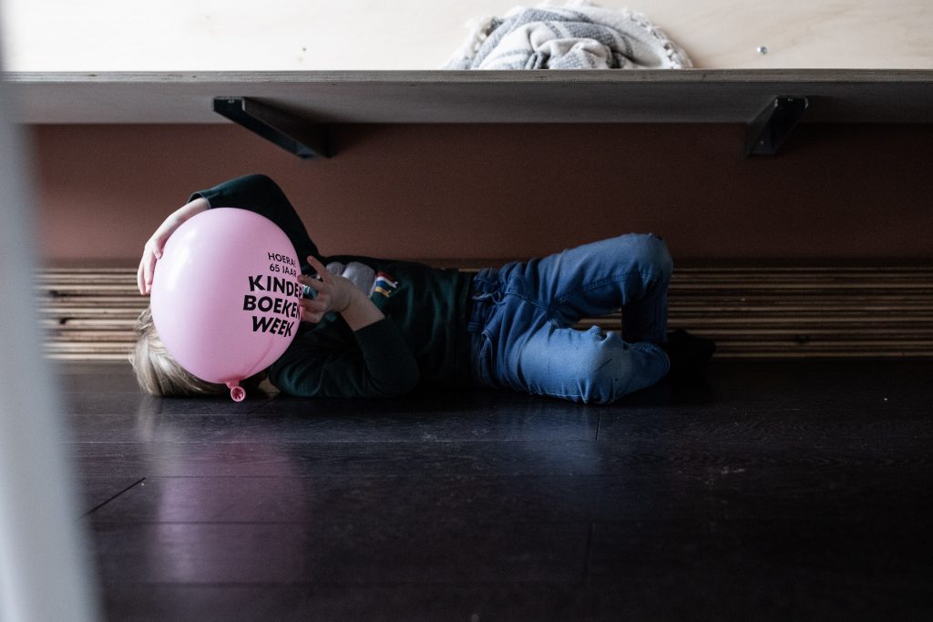 Day in the life, jongen ligt op de grond onder de bank en houdt een roze ballon voor zijn gezicht.