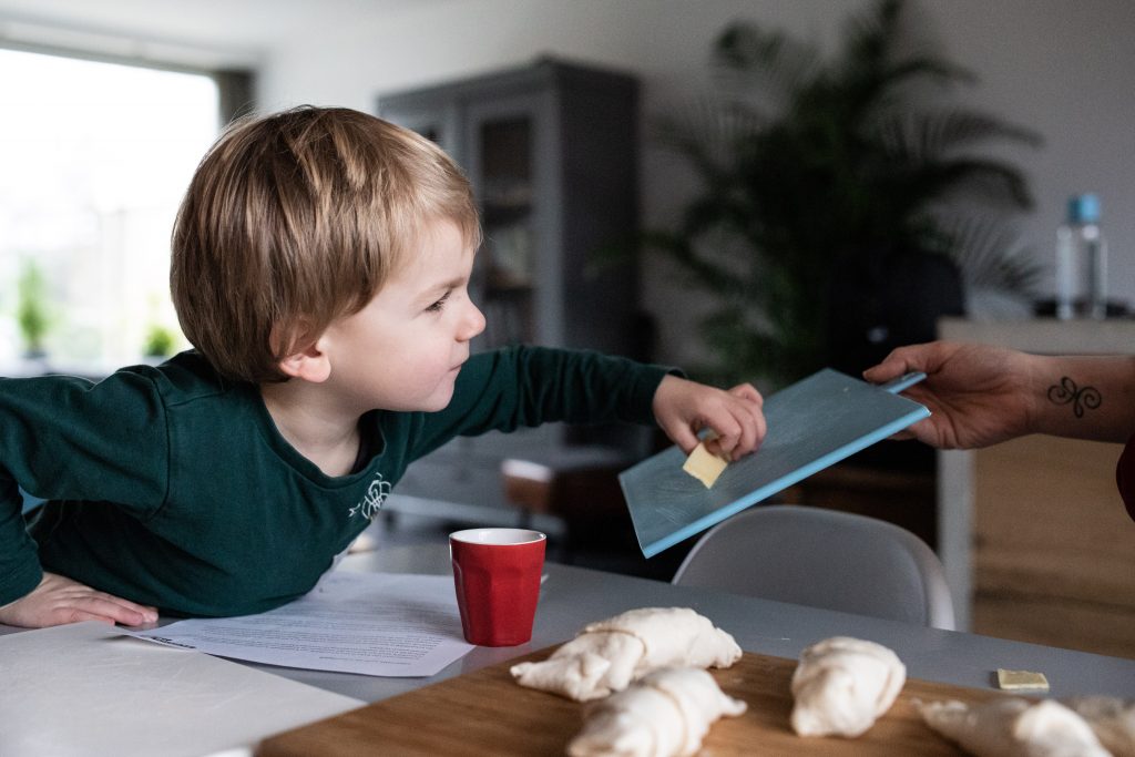 Een 'day in the life' met de familie Schoon, jongen buigt over de tafel heen om een plakje kaas van de plank te pakken.